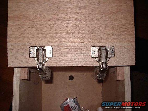 Two cabinet hinges attached to a wooden panel above an open box. A tape measure is visible inside the box