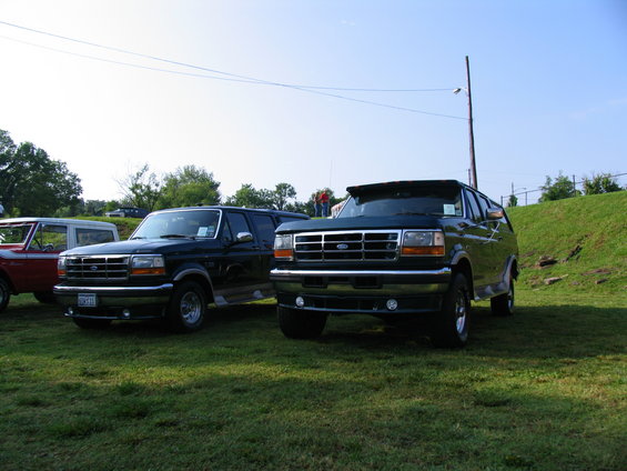 Two Ford Bronco SUVs parked on grass, one dark green, one black. A red vehicle is partially visible on the left