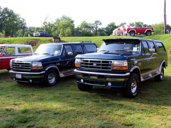 Two dark green Ford Bronco SUVs parked on grass, one in front of the other. A red pickup truck is visible on a hill in the background