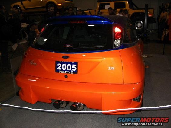 Rear view of an orange hatchback with a blue stripe and custom body kit. License plate reads 2005 POLITO. Dual exhaust pipes visible