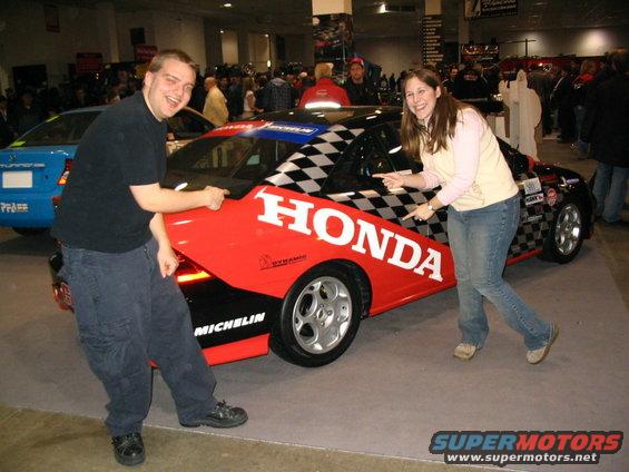 Guy and girl pointing at a red and black Honda race car with checkerboard pattern and "HONDA" text