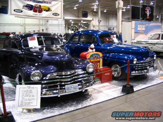 Dark purple and bright blue vintage sedans parked side-by-side indoors. A sign in front of the purple car reads '1949 CHEVY FLEETLINE'