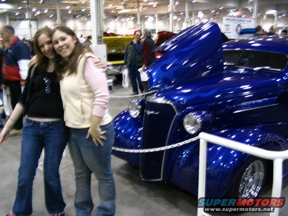 Two women pose in front of a shiny blue hot rod with flame graphics. The car's hood is open