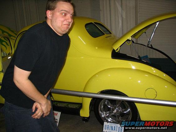 Guy in black shirt next to a yellow custom hot rod with flames on the side and hood open. License plate says URINATOR