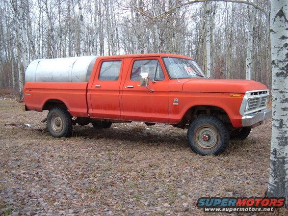 Orange crew cab pickup truck with a large silver tank in the bed, parked in a wooded area