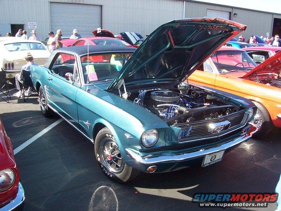 Teal 1965 Ford Mustang coupe with hood open, parked at a car show next to an orange car