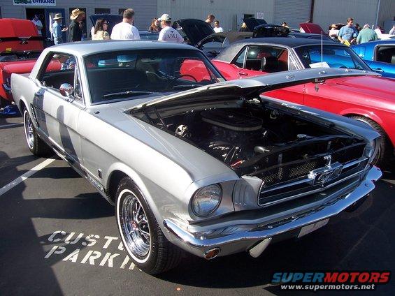 Silver 1965 Ford Mustang coupe with hood open, parked at an outdoor car show. Other cars visible in background