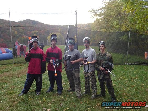 Five men in paintball gear stand in a grassy field with trees and a net in the background