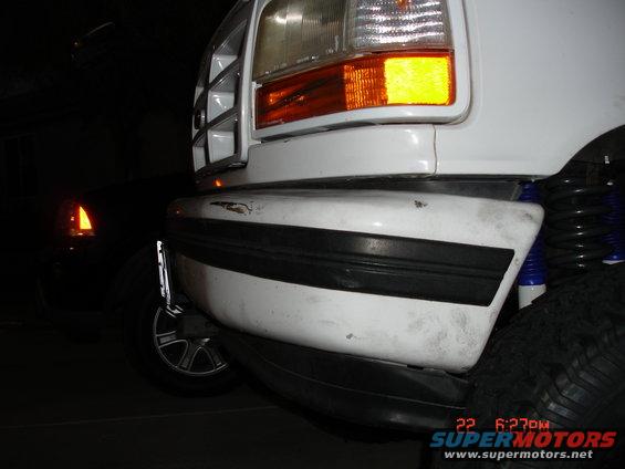 Front bumper and headlight of a white SUV, possibly a 1992 Ford Bronco. Another dark vehicle is visible in the background