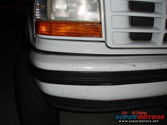 White Ford F-Series truck front bumper and headlight, showing a black rubber bumper trim and a portion of the tire