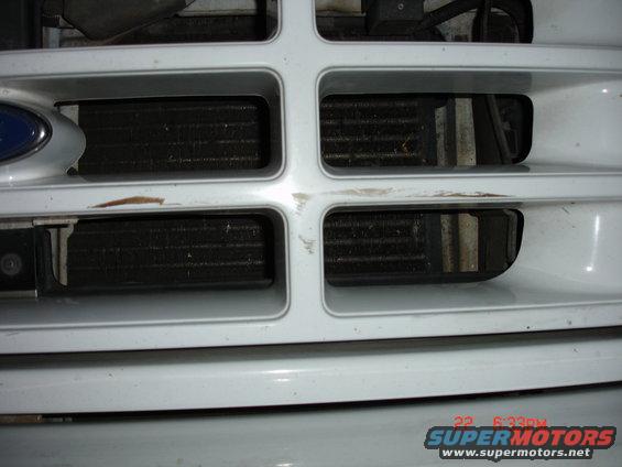 Close-up of a white Ford truck grille with two visible radiators behind it. The grille has a blue Ford emblem on the left