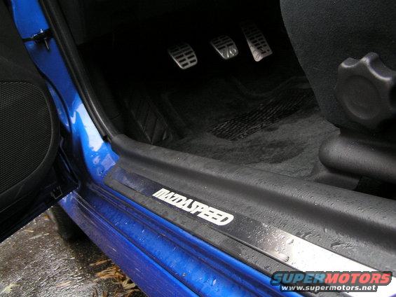 Blue car interior door sill with "MAZDASPEED" text, three pedals visible