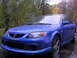 2003 Mazda Protege - A blue sedan is parked on a wet surface with trees in the background.