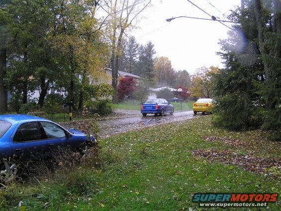 Blue sedan parked on grass, yellow and blue sedans driving on wet road