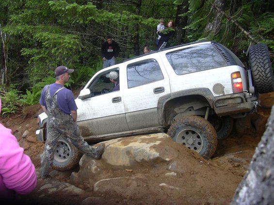 White SUV navigating a rocky, muddy trail with people watching