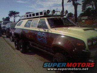 Custom six-wheeled SUV with a roof rack and lights. It has a brown and tan paint job with a blue logo on the side