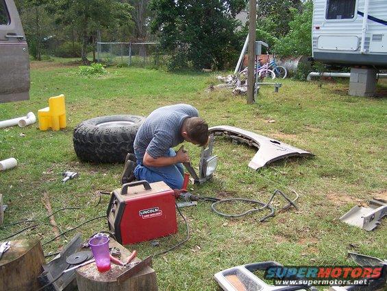 man welding a car fender and headlight assembly on grass, with a large tire and Lincoln welder nearby