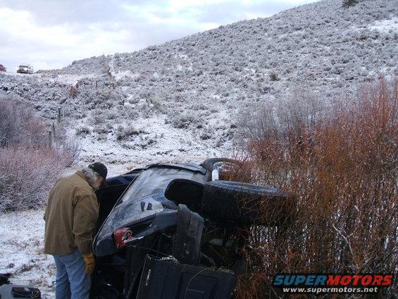 Pickup truck on its side in snowy brush, person looking at it. Another vehicle on hill