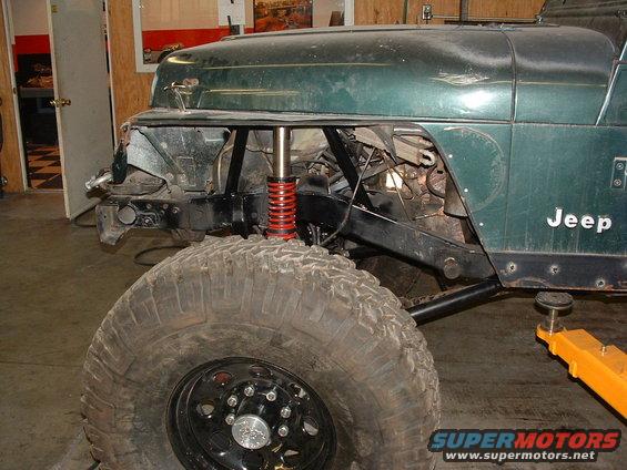 Green Jeep Wrangler with front end disassembled, showing suspension and engine components. Large off-road tire in foreground