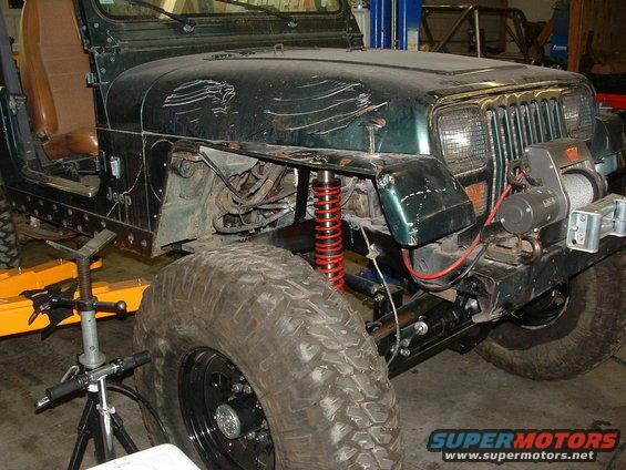 Green Jeep Wrangler with winch, lifted suspension, and large tires, showing significant body damage on the hood and fender