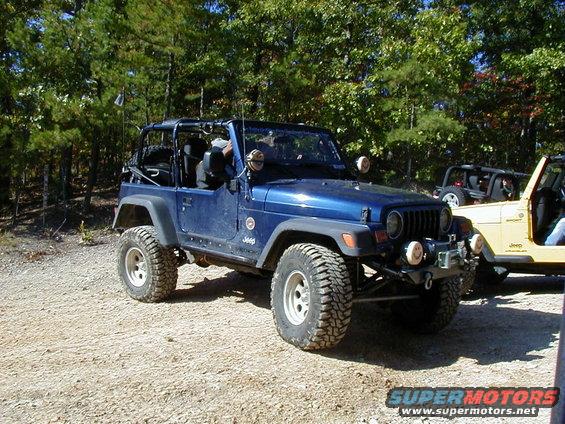 Blue Jeep Wrangler parked on a dirt road with trees in the background. Another Jeep is visible to the right