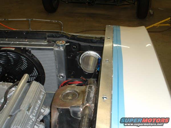 Car engine bay with radiator, fan, and part of the engine visible. White fender with blue stripe