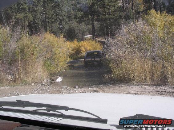 Dark SUV driving through a shallow stream in a wooded area with autumn foliage