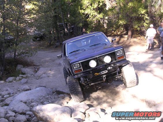 Dark blue off-road truck with large tires climbing over rocks on a dirt trail. Has two round headlights and a winch on the front