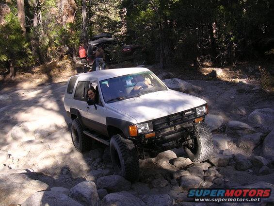 White 1985 Toyota 4Runner driving over rocks on a trail. Another vehicle is visible behind it