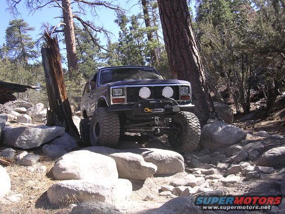Dark SUV with winch and off-road tires navigating rocky terrain. Appears to be a Ford Bronco from the 1980s