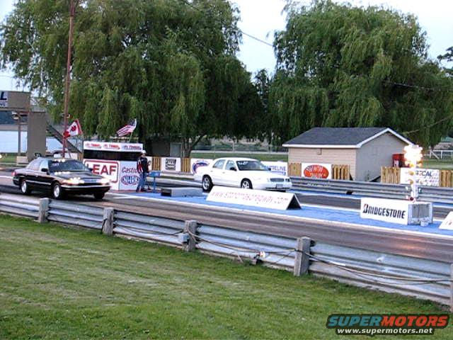 Two sedans, one black and one white, on a drag strip with trees and a small building in the background. The black car is in the foreground