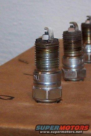 Three used spark plugs on a brown surface. The front plug shows wear on the electrode