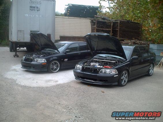 Two black sedans parked outside, hoods up showing engines. One has a strut bar