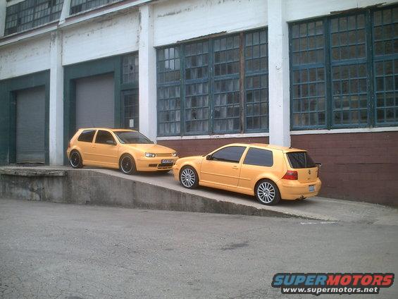 Two yellow Volkswagen Golf Mk4 hatchbacks parked on a ramp in front of an industrial building. The front car is facing left, the rear car is facing right
