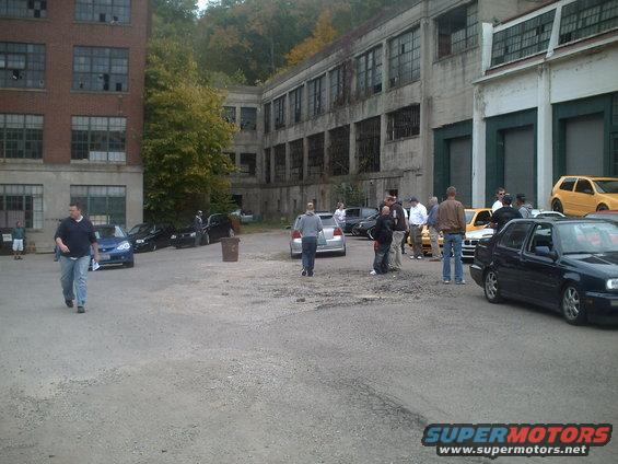 Several cars parked in a lot outside an old industrial building, with a group of people gathered