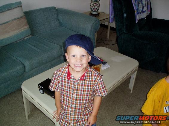 Child wearing a blue baseball cap and plaid shirt smiles at the camera, sitting next to a toy car on a coffee table