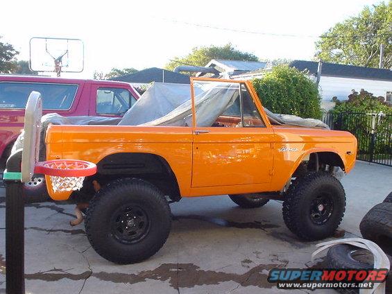 1977 Ford Bronco in orange, parked next to a red SUV, with a basketball hoop attached to the front fender