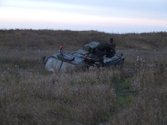 Military armored vehicle partially obscured by tall grass, with a fence in the background