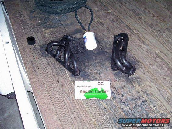 Two black exhaust headers, a tire, and a white canister with a hose on a wooden surface