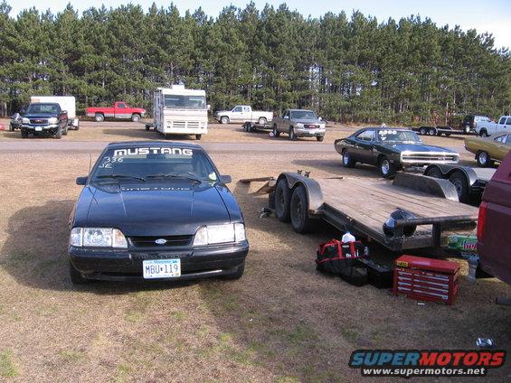 Front view of a black 1989 Ford Mustang with 'MUSTANG' decal on windshield and racing numbers. Trailer and other vehicles in background