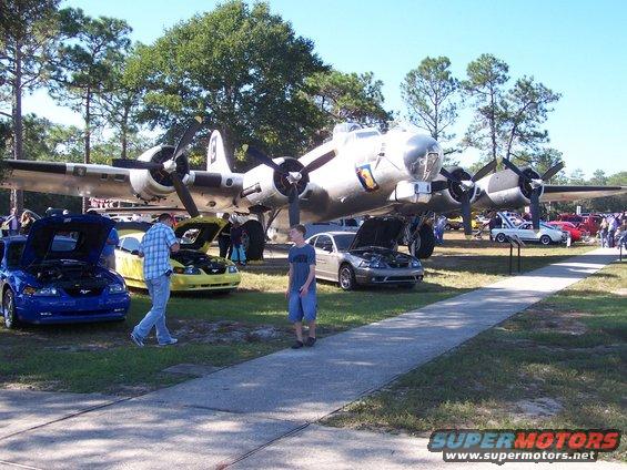 B-17 Flying Fortress airplane on display with several cars parked in front, some with hoods open