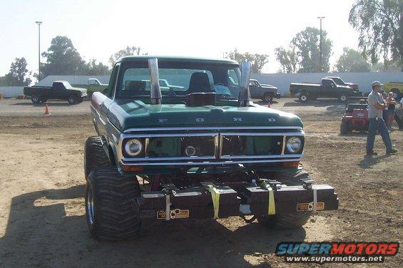 Green Ford pickup with huge tires and stacks. Other trucks and a person are in the background