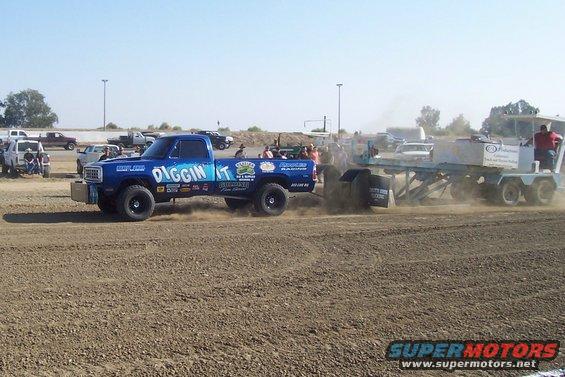 Blue pickup truck with "DIGGIN IT" graphics, pulling a sled in a dirt arena