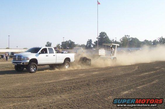 White Dodge Ram pickup truck pulling a sled in a dirt arena. The truck appears to be from the mid-2000s