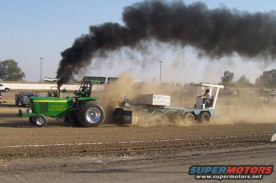 Green tractor with black smoke, pulling a sled on a dirt track