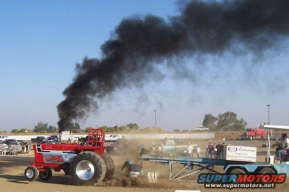 Red tractor with large rear wheels emitting black smoke during a pull
