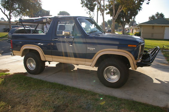 Blue 1986 Ford Bronco with tan trim and a black aftermarket bumper, parked on a driveway