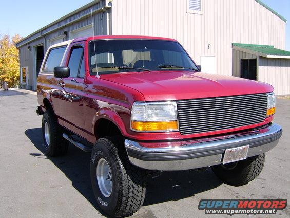 Red Ford Bronco with aftermarket grille parked outside a building. License plate is visible