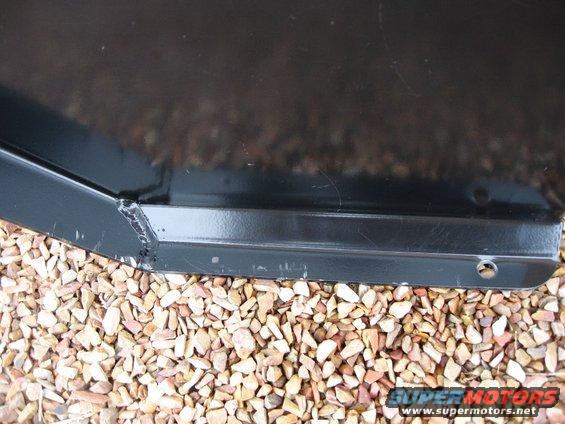black metal component with visible welds, resting on a bed of small stones