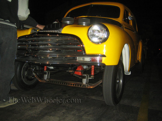Front view of a yellow hot rod with a large chrome grille and exposed engine components, including two air intakes. A person is standing by the open hood